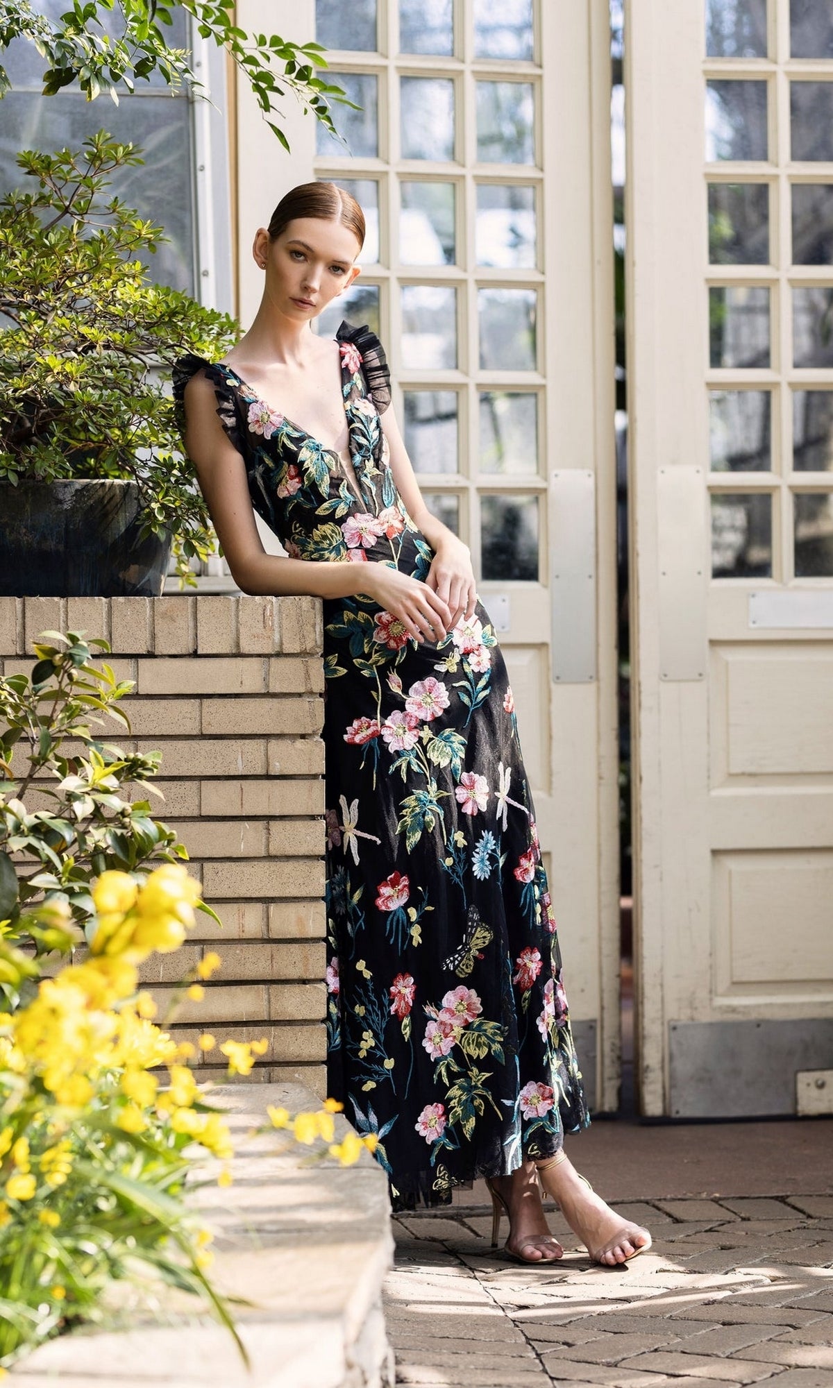 A woman wearing the Long Formal Dress 68273 by Mac Duggal, featuring black floral embroidery and a plunging v-neckline, stands by a brick wall and glass doors amid yellow flowers and greenery.